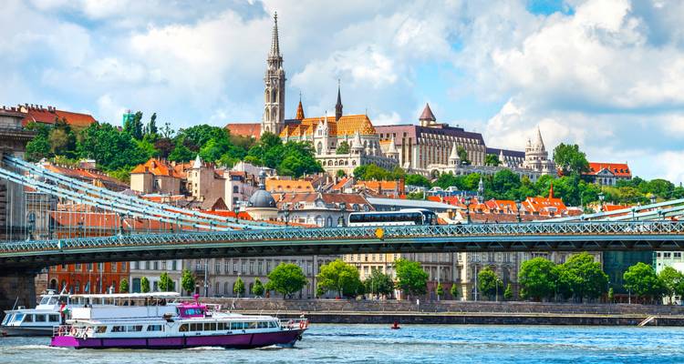 Un bateau de passagers passe sous le Pont des Chaînes de Budapest avec la Colline du Château de Buda et l'Église Matthias qui se dressent en arrière-plan.