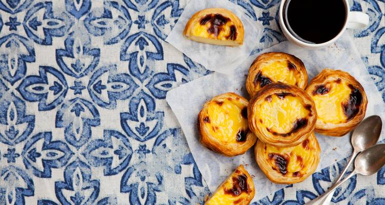 Tartas de crema portuguesa recién horneadas y café dispuestos sobre un mantel con estampado estilo azulejo.