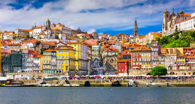 Las coloridas fachadas fluviales del distrito Ribeira de Oporto coronadas por iglesias históricas y una torre bajo un cielo azul.