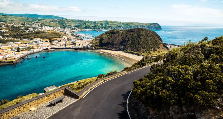 Carretera sinuosa con vistas a la bahía esmeralda y el pueblo costero de las Azores bajo cielos soleados.
