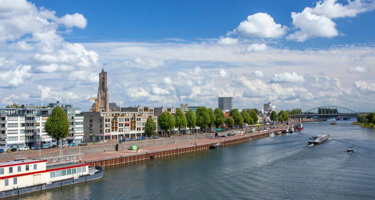 Un río bordeado de edificios modernos, árboles y botes amarrados se extiende bajo un cielo de nubes blancas y esponjosas.