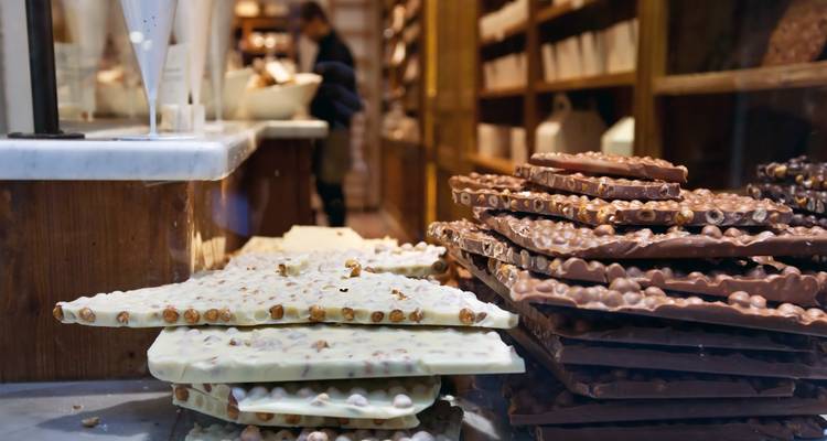 Stapel von handwerklich gefertigter Milch- und Weißschokolade mit Haselnüssen in einer Boutique-Chocolaterie, unscharfer Chocolatier im Hintergrund.