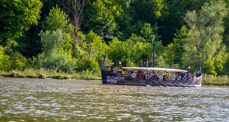 Bateau viking en bois nommé Neufundland naviguant sur une rivière calme avec une forêt verte luxuriante.