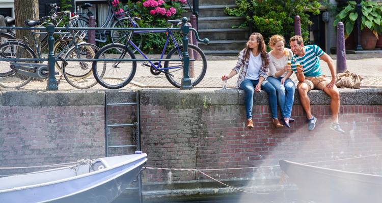 Tres amigos colgando sus piernas sobre el borde de un canal de Ámsterdam disfrutando de bebidas con bicicletas estacionadas atrás.