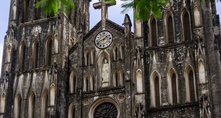 Gros plan de la façade usée de la cathédrale Saint-Joseph de Hanoï avec horloge centrale et détails gothiques partiellement encadrés par des feuilles vertes.