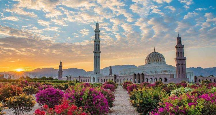 La Grande Mosquée du Sultan Qaboos encadrée par des jardins en fleurs au lever du soleil avec des montagnes au-delà.