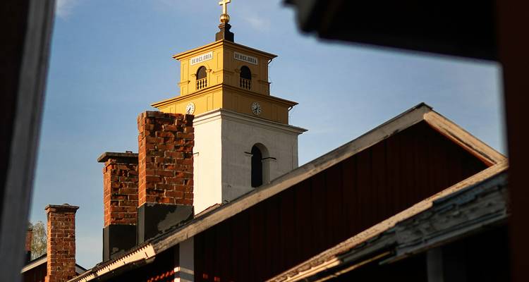 Sun-lit yellow church tower with brick chimneys and wooden rooftops framed against a blue sky.