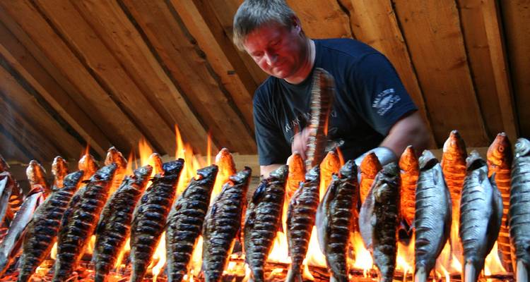 Chef grills rows of whole fish over open flames inside a wooden smokehouse.