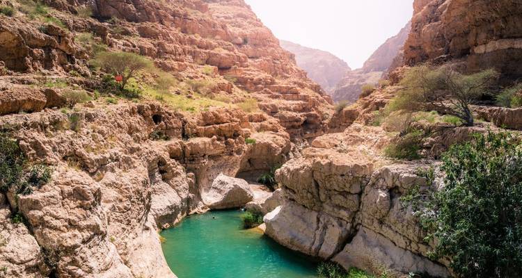 Piscine turquoise nichée au sein de parois de canyon en pierre rouge escarpée à Wadi Shab avec un randonneur solitaire sur la falaise.