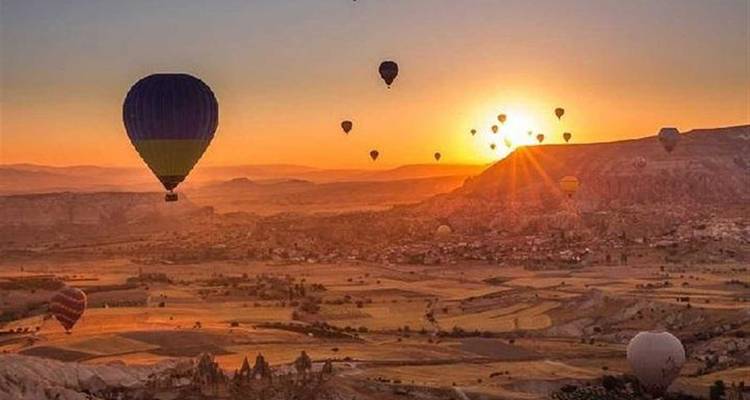 Farbenfrohe Heißluftballons schweben bei Sonnenaufgang über ein breites Tal und werfen lange Schatten auf die Flickenteppich-Landschaft.