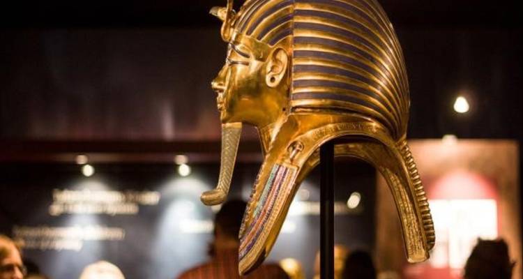 Goldene Totenmaske des Tutanchamun, die unter dramatischer Beleuchtung in einer Museumsgalerie ausgestellt ist.