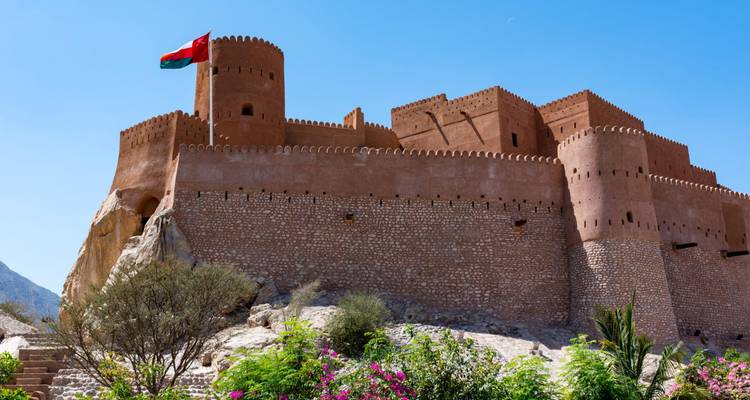 Fuerte de Nakhal bañado por el sol con la bandera omaní ondeando alto sobre muros defensivos escalonados y plantas desérticas.