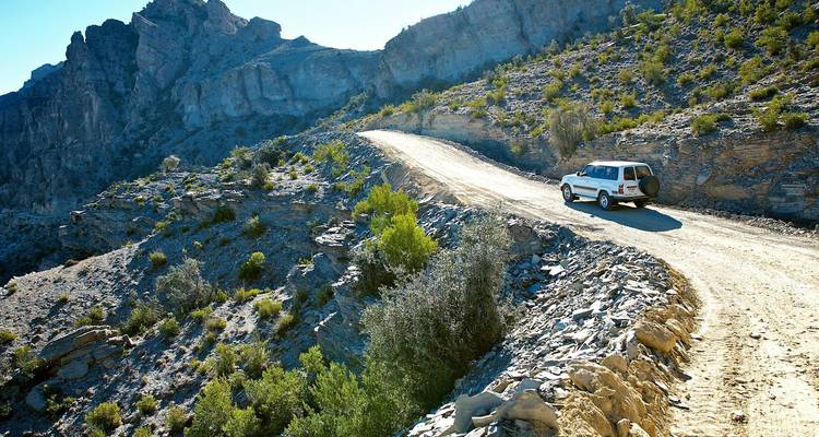Vehículo 4x4 conduciendo por un sinuoso camino de montaña de grava con acantilados rocosos asomándose a su lado.