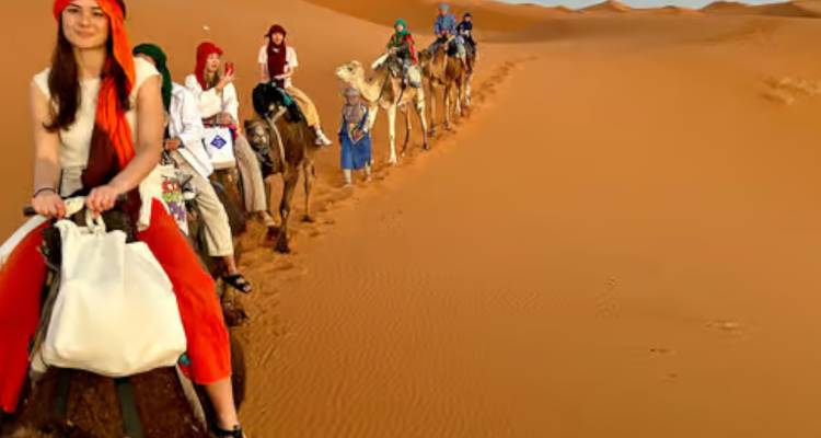 Une file de voyageurs montés sur des chameaux serpente à travers les dunes ouvertes, menée par une femme en tête.