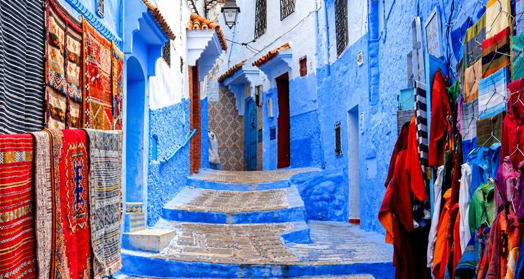 Alfombras y telas coloridas bordeando un callejón azul con escalones en Chefchaouen.