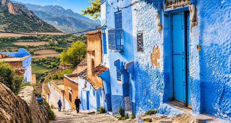 Dos personas caminando por una calle de piedra pasando casas azules con vistas a un valle fértil.