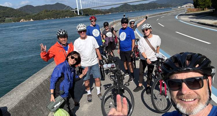 Radfahrer versammeln sich auf einer Küstenstraße neben dem blauen Meer und einer Hängebrücke und winken fröhlich.