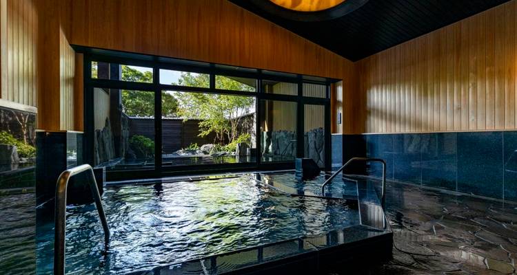 Ruhiges Innen-Onsen-Becken mit Steinboden und großem Fenster mit Blick auf einen Garten.