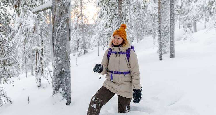 A smiling woman in winter gear stands knee-deep in fresh snow among snow-laden pines in soft morning light.