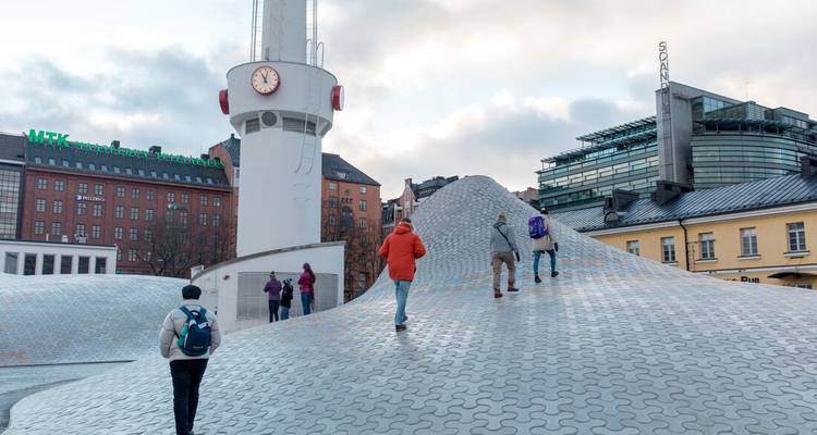 People explore a futuristic wavy plaza of light-gray tiles beside a clock tower in a Nordic city.