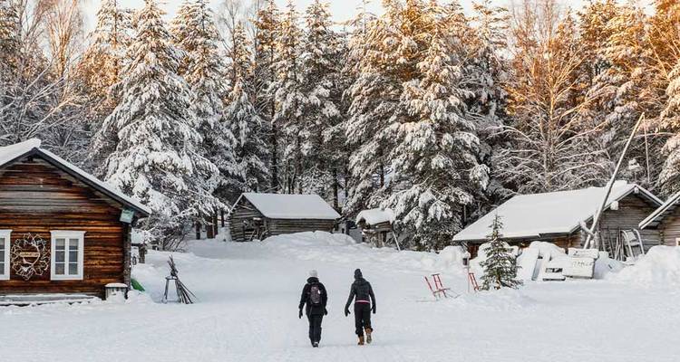 Two travelers walk toward rustic log cabins surrounded by tall snow-covered pines glowing in winter light.