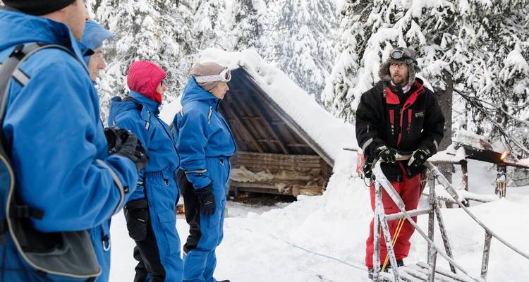 A guide explains gear to travelers in blue winter suits beside a snowy shelter and campfire.