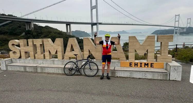 Radfahrer feiert mit erhobenen Armen vor dem hölzernen Shimanami-Schild und der Brücke im Hintergrund.