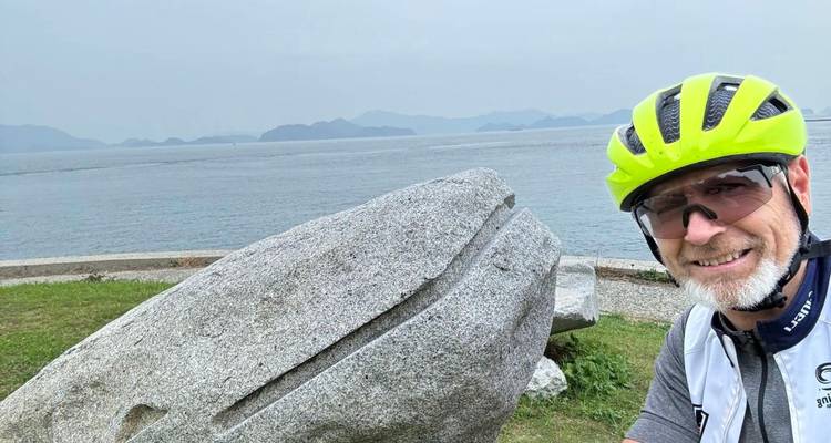 Ein Radfahrer in einem leuchtend farbigen Helm lächelt neben einer großen Steinskulptur auf einem Küstenpfad.
