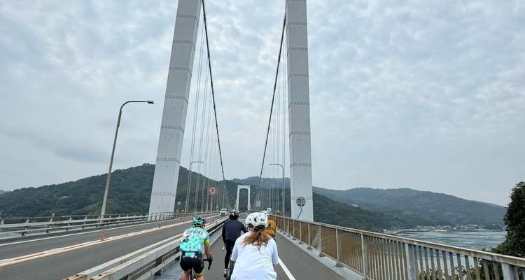 Radfahrer fahren über eine hohe Hängebrücke unter teilweise bewölktem Himmel und Hügeln.