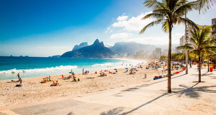 Vue ensoleillée de la promenade de la plage d'Ipanema bordée de palmiers et de montagnes lointaines.
