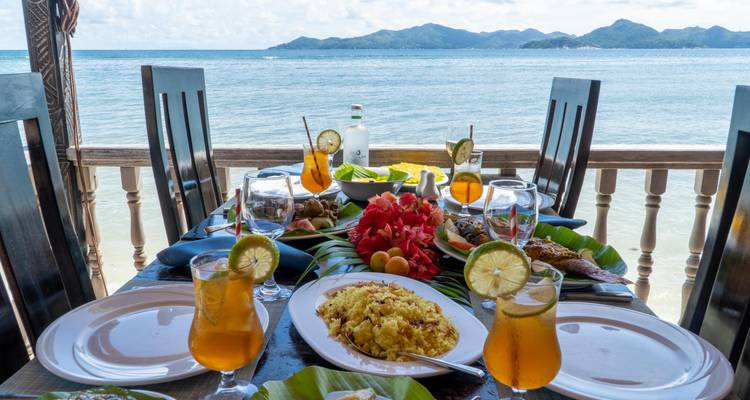 Table en front de mer chargée de plats tropicaux, de fruits et de boissons glacées surplombant une mer bleu clair et des îles vertes lointaines.