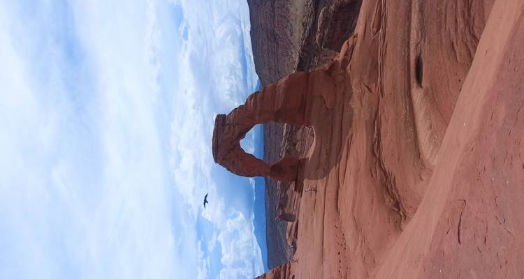 Iconic Delicate Arch standing on a red sandstone ridge with tiny hikers below under a partly cloudy sky.