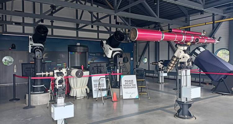 Indoor observatory exhibit with multiple large telescopes behind ropes and informational signs.