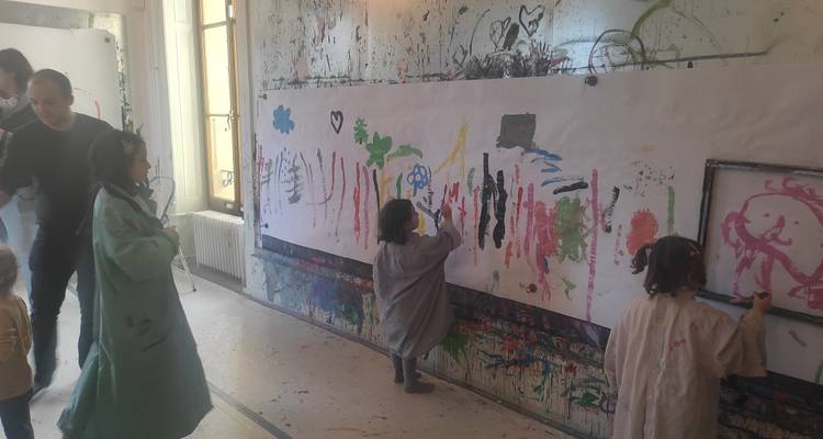 Des enfants en blouses de peinture créent avec enthousiasme une œuvre d'art sur une longue fresque en papier à l'intérieur d'un studio désordonnés.