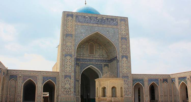Innenhofblick einer aufwendig gekachelten Madrasa mit großem Bogenportal und blauer Kuppel unter dunstigem Himmel.