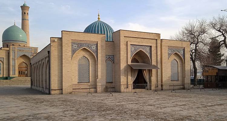 Komplexe Backsteinmoschee mit türkisfarbenen Kuppeln und Minarett, die einen geräumigen Steinhof begrenzt.