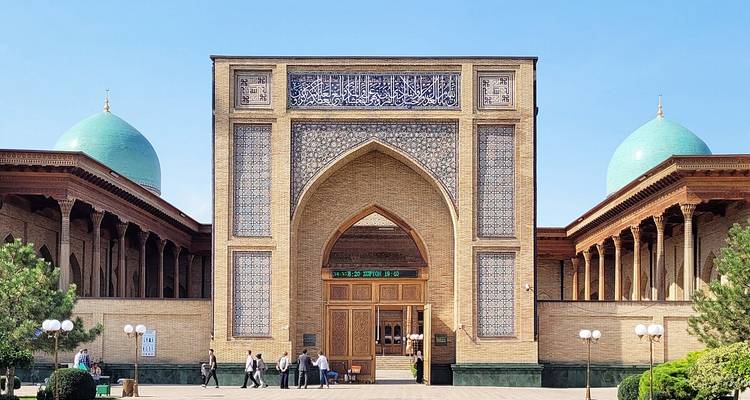 Vorderfassade einer islamischen Madrasa mit aufragendem Iwan und zwei türkisfarbenen Kuppeln, Besucher darunter.