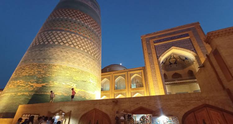 Abendaufnahme des beleuchteten Kachelturms und der Madrasa-Mauern in warmem goldenen Licht mit Besuchern darunter.
