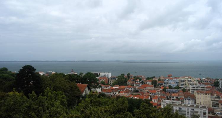 Panoramablick über eine rotgedeckte Hafenstadt zum weitläufigen Buchtgewässer unter bedecktem Himmel.