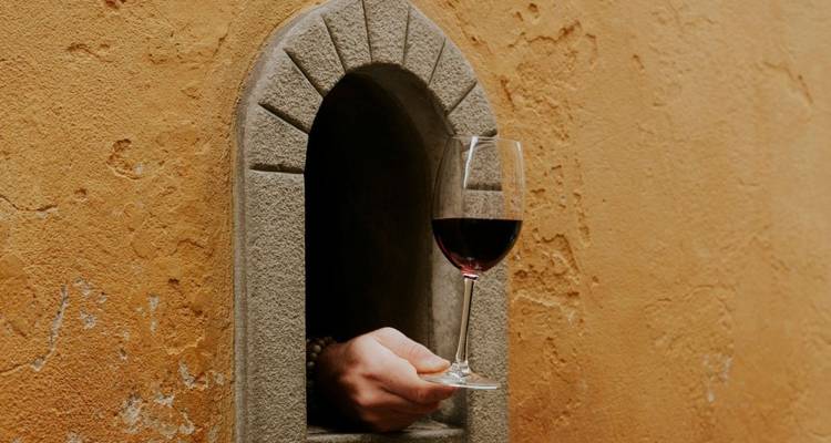 Una mano pasando una copa de vino tinto a través de una pequeña abertura de piedra con arco en una pared ocre.