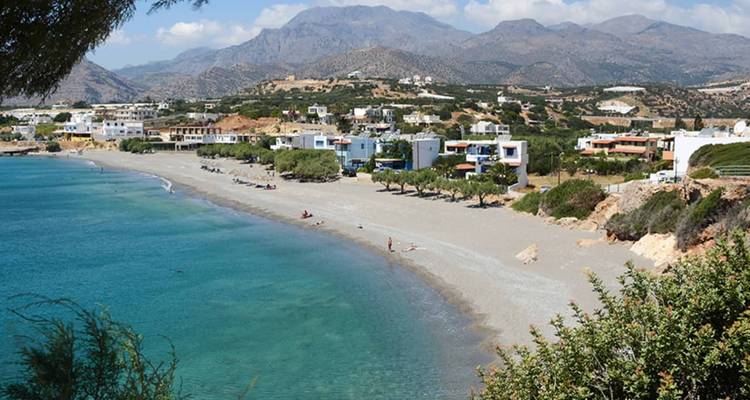 Breiter Sandstrand mit klarem türkisfarbenem Wasser und einem kleinen Küstendorf, das von rauen Bergen umgeben ist.