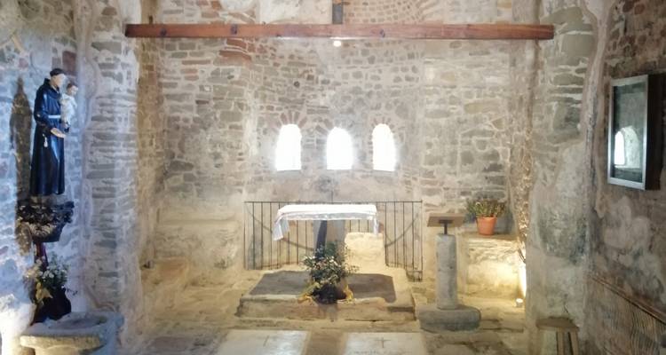Interior de una pequeña capilla de piedra con un altar sencillo, ventanas arqueadas y una estatua religiosa en luz suave