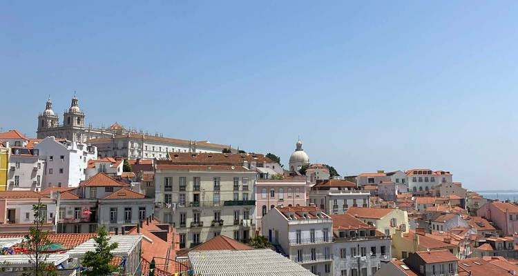 Panoramic view over Alfama’s orange-tiled rooftops with church domes and the River Tagus in the background.