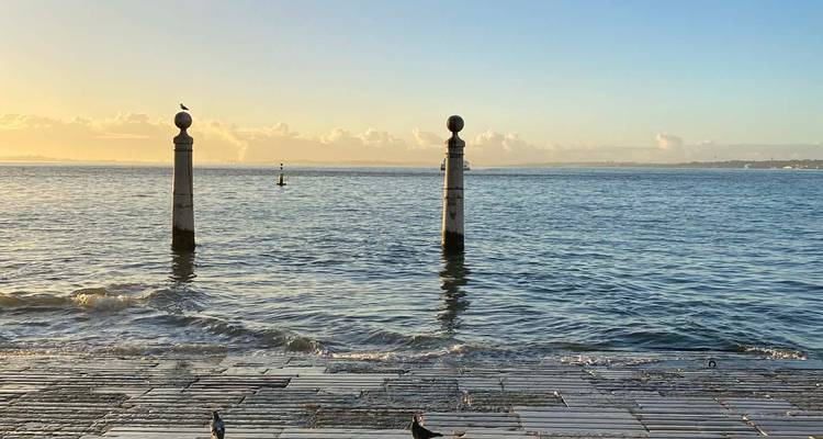 Two stone columns stand in shallow water of the Tagus River at Cais das Colunas during golden sunrise.