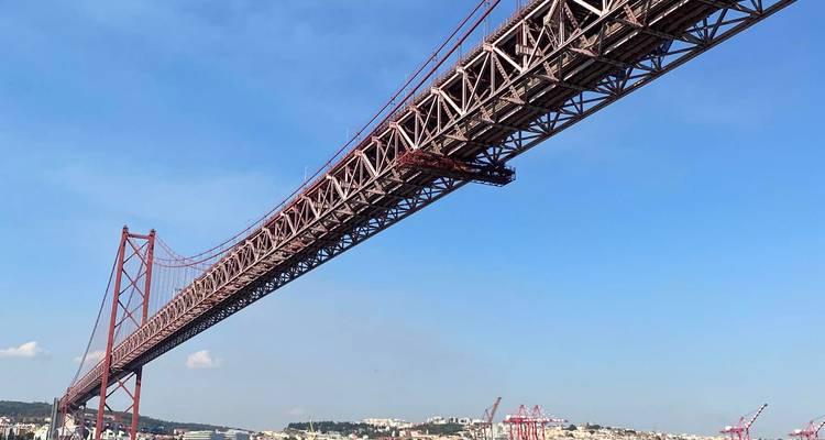 Long red suspension bridge stretching across a wide river under a clear blue sky with city buildings in the distance.