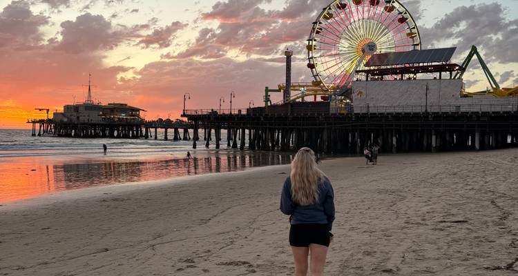 Un visiteur marchant le long de la plage de Santa Monica au coucher du soleil avec la jetée et la grande roue illuminées.