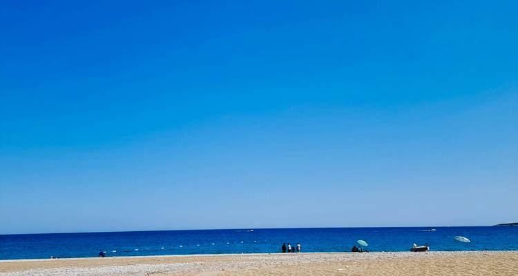 Playa ancha de arena y grava con aguas turquesas del Mediterráneo y unos pocos bañistas bajo sombrillas.