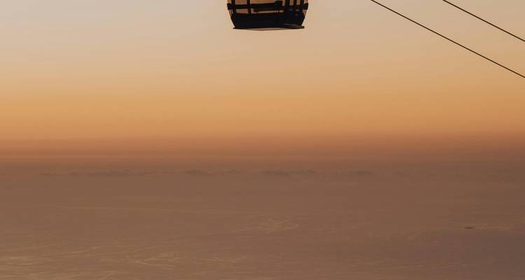 Silueta de una cabina de teleférico suspendida contra un cielo naranja al atardecer sobre el mar.