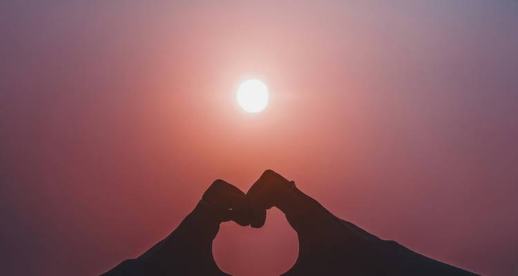 Twee handen vormen een hartje dat de zon omlijst in een mistige roze lucht.