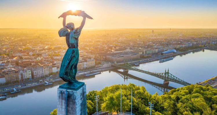 Estatua de la Libertad sosteniendo una corona de laurel sobre el panorama iluminado por el amanecer de Budapest y el Danubio